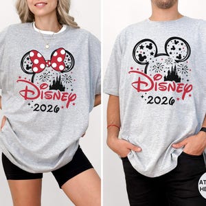 Puede incluir: Una camiseta gris claro con una silueta negra de las orejas de Mickey Mouse rellena de fuegos artificiales y estrellas. El dise&ntilde;o incluye el logo de Disney en rojo, el a&ntilde;o 2026 en negro y un lazo rojo de lunares en la versi&oacute;n izquierda.