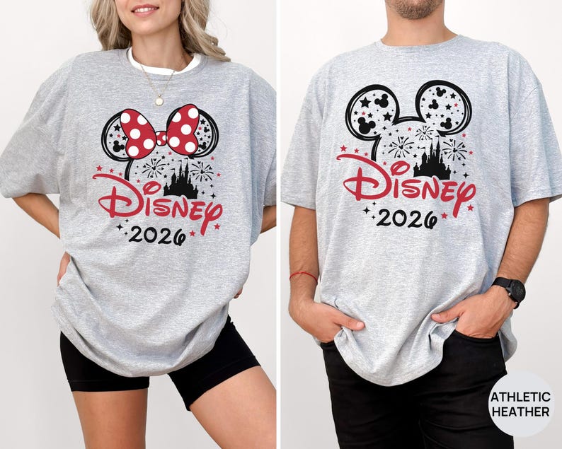 Puede incluir: Una camiseta gris claro con una silueta negra de las orejas de Mickey Mouse rellena de fuegos artificiales y estrellas. El dise&ntilde;o incluye el logo de Disney en rojo, el a&ntilde;o 2026 en negro y un lazo rojo de lunares en la versi&oacute;n izquierda.