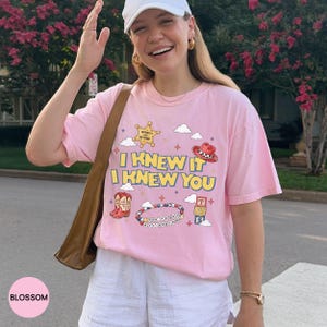 Op de afbeelding: Een lichtroze t-shirt met de gele tekst I KNEW IT I KNEW YOU, omringd door afbeeldingen van een sheriffster, een cowboyhoed, een westernlaars, een kralenarmband met tekst, alfabetblokken en kleine wolkjes en sterretjes.