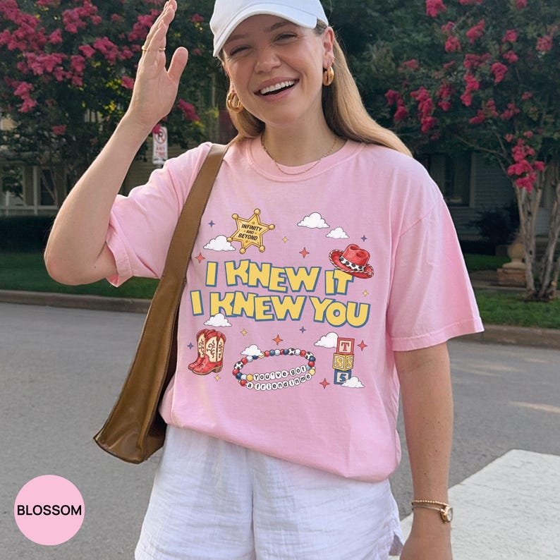 Op de afbeelding: Een lichtroze t-shirt met de gele tekst I KNEW IT I KNEW YOU, omringd door afbeeldingen van een sheriffster, een cowboyhoed, een westernlaars, een kralenarmband met tekst, alfabetblokken en kleine wolkjes en sterretjes.