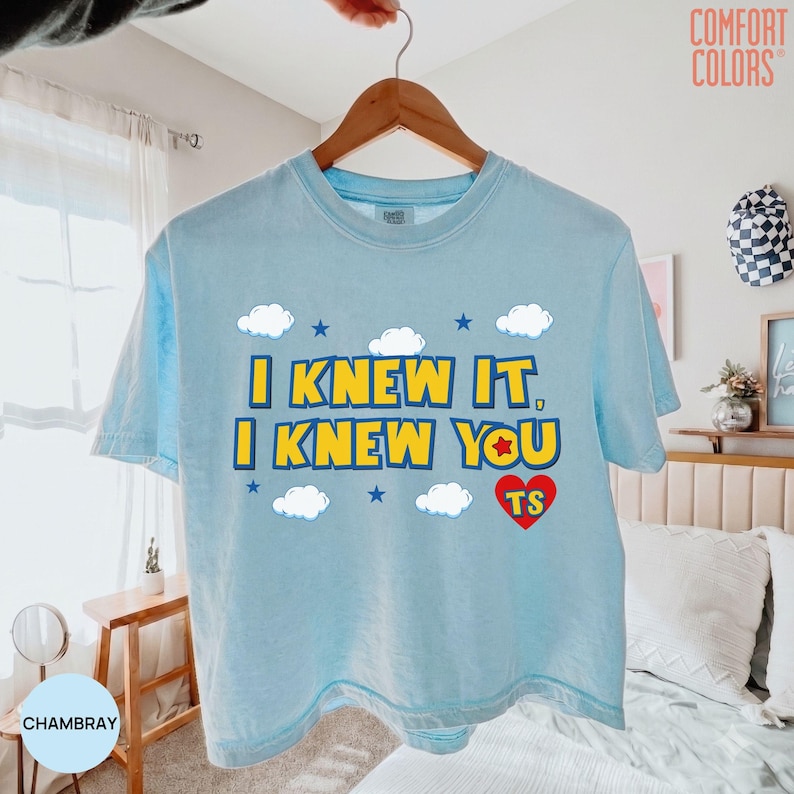May include: A light blue short-sleeve t-shirt featuring the yellow text I KNEW IT, I KNEW YOU with a red heart containing the letters TS, surrounded by white clouds and small blue stars. The shirt is displayed on a wooden hanger.