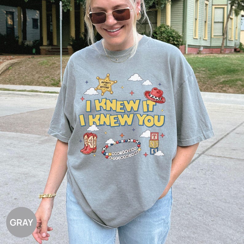 Op de afbeelding: Een grijs t-shirt met korte mouwen en een grafische print met de tekst I KNEW IT I KNEW YOU. Het ontwerp bevat een gele sheriffster, een rode cowboyhoed, rode cowboylaarzen, een kralenarmband en alfabetblokken met wolken en kleine sterren.