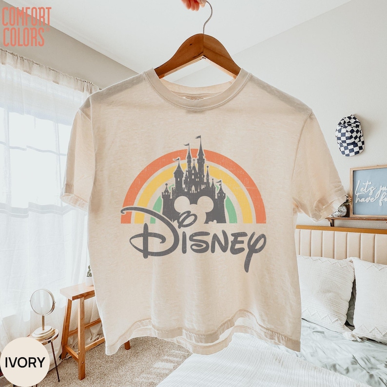 May include: An ivory short-sleeve t-shirt featuring a graphic print of a gray castle silhouette centered in front of a multi-colored rainbow arch. The word Disney is printed in a gray script font below the castle. The shirt has a crew neckline.