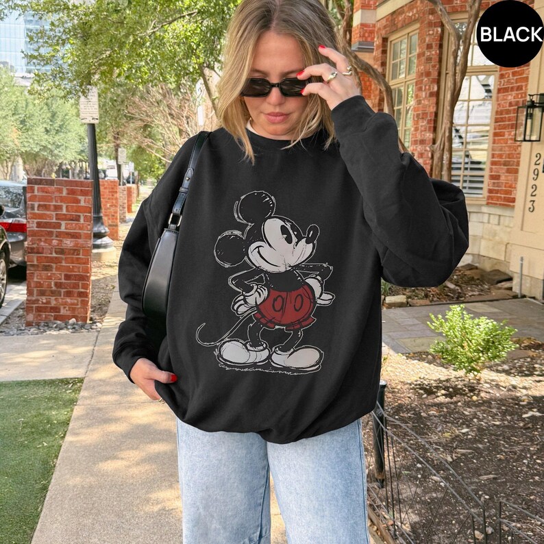 May include: Black sweatshirt featuring a distressed graphic of Mickey Mouse in a classic pose. The cartoon character is wearing red shorts and white gloves. The sweatshirt has a crew neck and long sleeves.