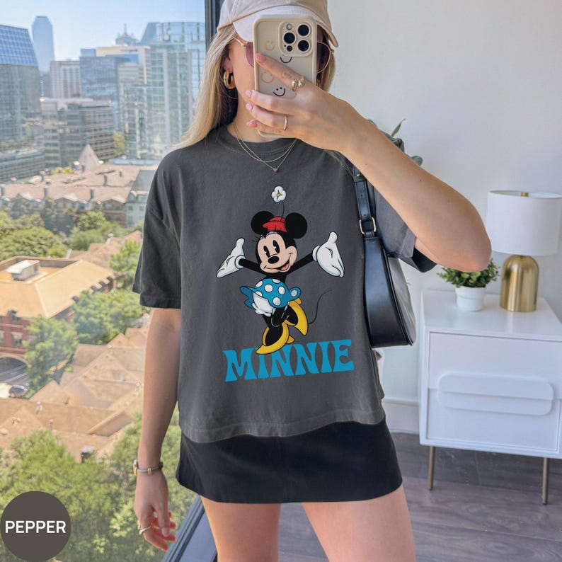May include: A charcoal gray short-sleeve t-shirt featuring a graphic print of Minnie Mouse wearing a red hat and yellow shoes, holding a blue and white polka-dot umbrella. The name MINNIE is printed in large, bold, light blue capital letters below.