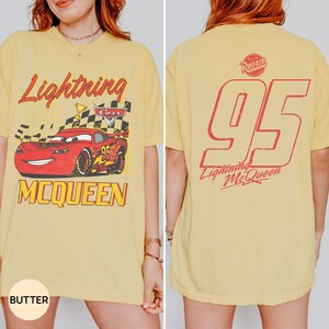 May include: Butter-colored t-shirt featuring a graphic of Lightning McQueen from the Cars movie. The front displays the car with text "Lightning McQueen" and "Cars". The back shows a large "95" and "Lightning McQueen" in red.