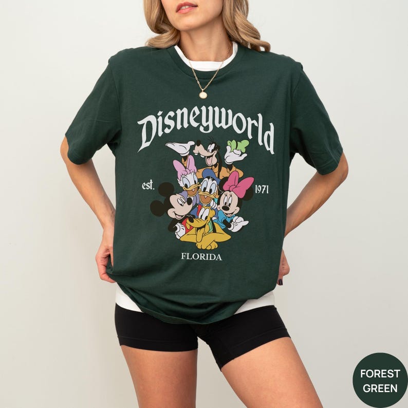May include: A forest green short-sleeve t-shirt featuring a white graphic print that reads Disneyworld, est. 1971, and FLORIDA. The design includes colorful illustrations of Mickey Mouse, Minnie Mouse, Donald Duck, Daisy Duck, Goofy, and Pluto.