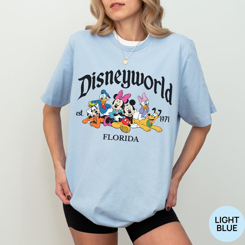 May include: A light blue short-sleeve t-shirt featuring a graphic of Disney characters including Mickey Mouse, Minnie Mouse, Donald Duck, Daisy Duck, Goofy, and Pluto. The text reads Disneyworld est. 1971 FLORIDA in black lettering.