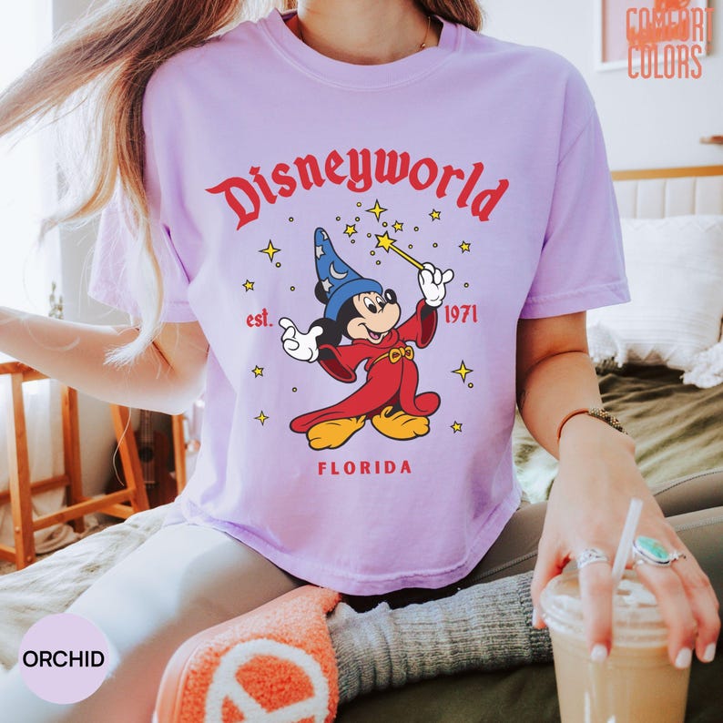 May include: A light purple t-shirt with a Disney World graphic. The graphic features Mickey Mouse in a wizard outfit, holding a wand, with the words "Disneyworld" and "Florida" in red. The shirt also has the text "est. 1971".