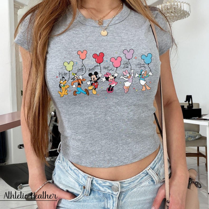 May include: Gray crop top with a graphic featuring Disney characters holding balloon-shaped balloons. The characters include Pluto, Goofy, Mickey Mouse, Minnie Mouse, Daisy Duck, and Donald Duck. The text "Athletic Heather" is printed at the bottom.