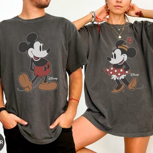 May include: Two charcoal gray t-shirts featuring Mickey and Minnie Mouse. Mickey is wearing red shorts and brown shoes. Minnie is wearing a red polka dot skirt and brown shoes. Both shirts have the Disney logo.