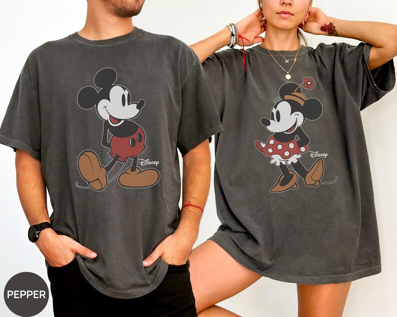 May include: Two charcoal gray t-shirts featuring Mickey and Minnie Mouse. Mickey is wearing red shorts and brown shoes. Minnie is wearing a red polka dot skirt and brown shoes. Both shirts have the Disney logo.