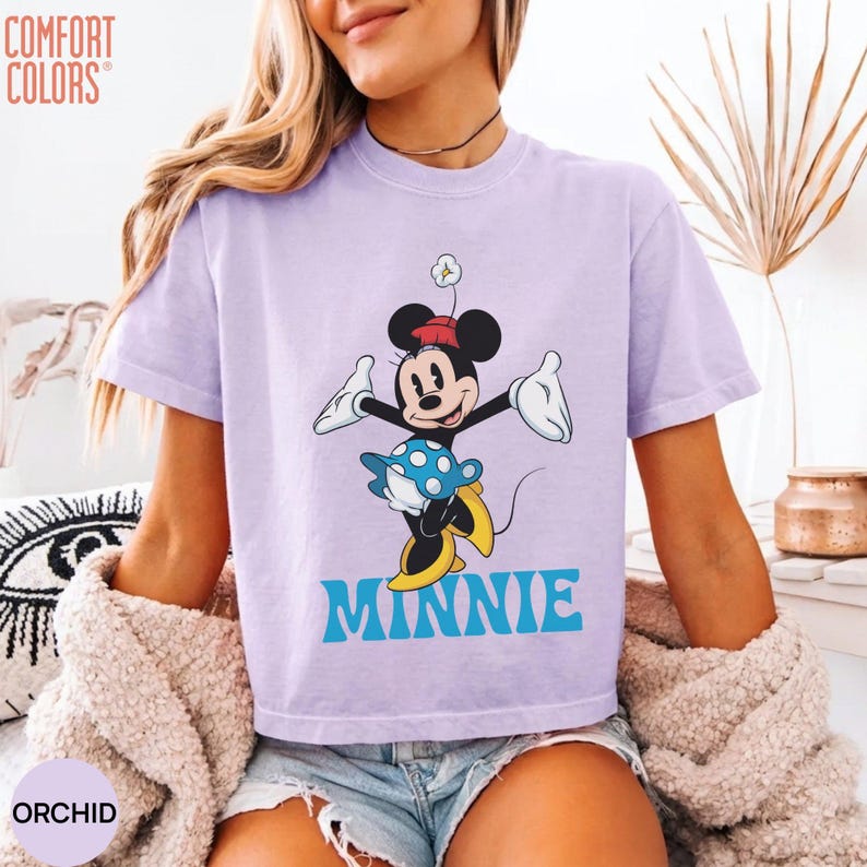 May include: A light purple short-sleeve t-shirt featuring a graphic of Minnie Mouse wearing a red hat and a blue polka-dot skirt. The character is depicted with white gloves and yellow shoes. The word MINNIE is printed in large blue letters below.