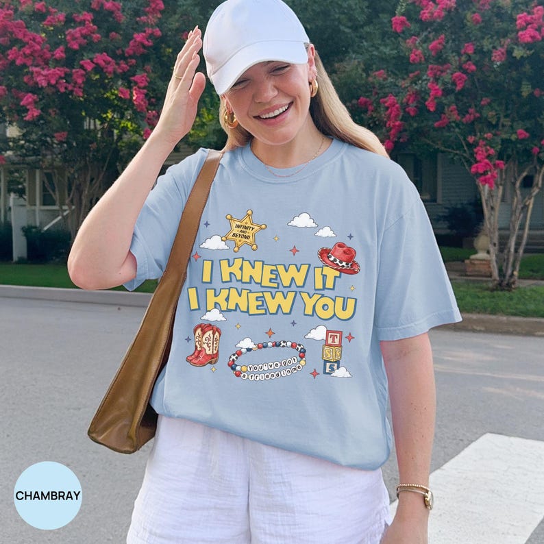 Op de afbeelding: Een lichtblauw t-shirt met een grafische print met de tekst I KNEW IT I KNEW YOU, een sheriffster, een rode cowboyhoed, cowboylaarzen, een kralenarmband met de tekst YOU'VE GOT A FRIEND IN ME en speelgoedblokken met letters.