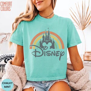 May include: A mint green short-sleeve t-shirt featuring a graphic of a gray castle silhouette centered in front of a multi-colored pastel rainbow. The word Disney is printed in a gray script font below the castle. The text Comfort Colors is visible.