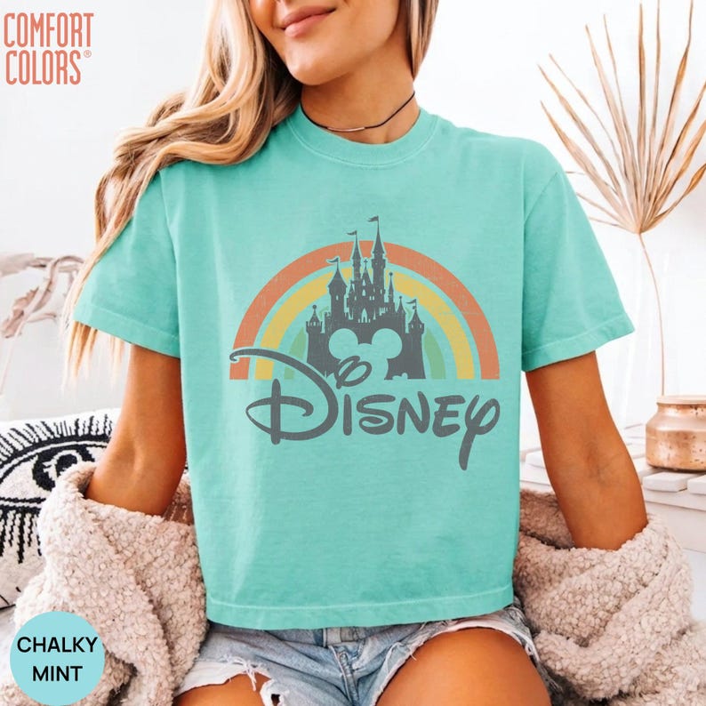 May include: A mint green short-sleeve t-shirt featuring a graphic of a gray castle silhouette centered in front of a multi-colored pastel rainbow. The word Disney is printed in a gray script font below the castle. The text Comfort Colors is visible.
