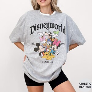 May include: An athletic heather gray t-shirt featuring the black text Disneyworld est. 1971 Florida. The graphic includes a colorful illustration of Mickey Mouse, Minnie Mouse, Donald Duck, Daisy Duck, Goofy, and Pluto grouped together in the center.