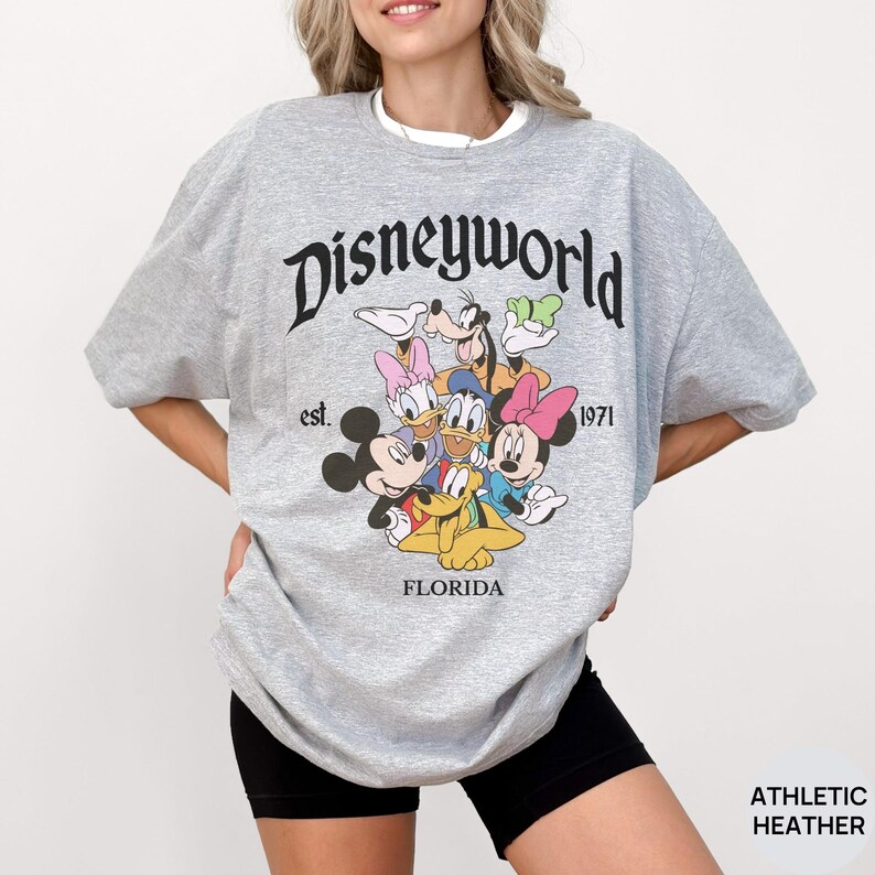 May include: An athletic heather gray t-shirt featuring the black text Disneyworld est. 1971 Florida. The graphic includes a colorful illustration of Mickey Mouse, Minnie Mouse, Donald Duck, Daisy Duck, Goofy, and Pluto grouped together in the center.