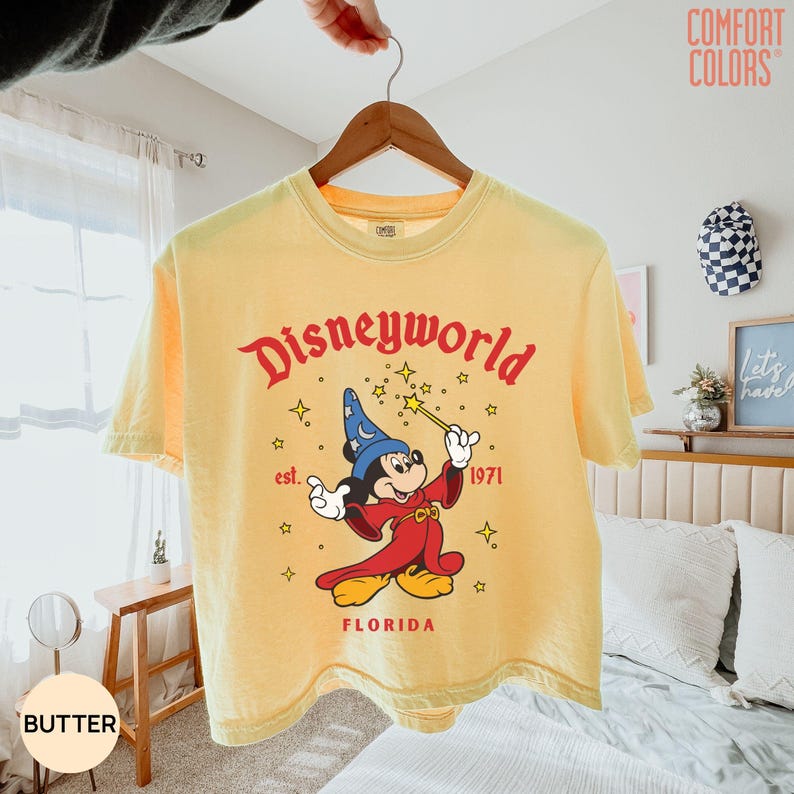 May include: Butter-colored t-shirt with a graphic of Mickey Mouse as a sorcerer, holding a wand. The shirt reads "Disneyworld" in red script, with "est. 1971" and "FLORIDA" below. The shirt is hanging on a wooden hanger.