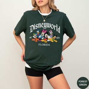 May include: A forest green short-sleeve t-shirt featuring a graphic print of Disney characters including Mickey Mouse, Minnie Mouse, Donald Duck, Daisy Duck, Goofy, and Pluto. The text reads Disneyworld est. 1971 FLORIDA in white lettering.