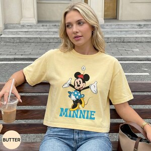 May include: A pale yellow short-sleeve t-shirt featuring a graphic of Minnie Mouse wearing a blue polka-dot skirt and yellow shoes. The character is depicted with arms outstretched, and the word MINNIE is printed in large blue capital letters below.