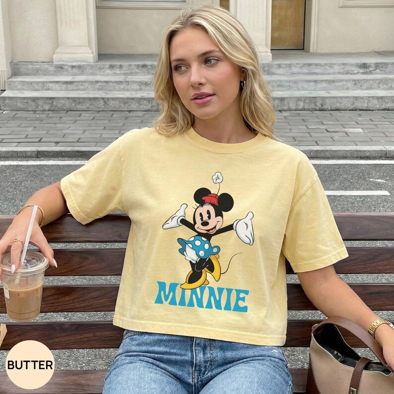 May include: A pale yellow short-sleeve t-shirt featuring a graphic of Minnie Mouse wearing a blue polka-dot skirt and yellow shoes. The character is depicted with arms outstretched, and the word MINNIE is printed in large blue capital letters below.