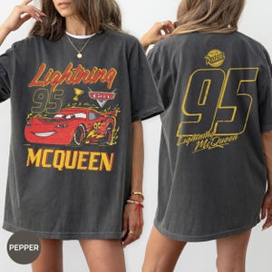 May include: Dark gray t-shirt featuring a graphic of Lightning McQueen from the Cars movie. The front design includes the text "Lightning 95 McQueen" with a red car illustration. The back displays a large "95" and "Lightning McQueen" in yellow.