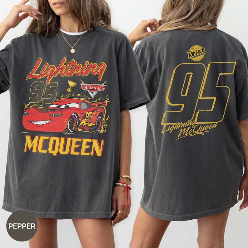 May include: Dark gray t-shirt featuring a graphic of Lightning McQueen from the Cars movie. The front design includes the text "Lightning 95 McQueen" with a red car illustration. The back displays a large "95" and "Lightning McQueen" in yellow.