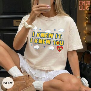 May include: An ivory short-sleeve t-shirt featuring the blue text I KNEW IT, I KNEW YOU with a red heart containing the letters TS, surrounded by small blue stars and white clouds. The shirt has a crew neckline and a relaxed fit design.