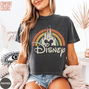 May include: A charcoal gray short-sleeve t-shirt featuring a graphic print of a white castle silhouette centered under a multi-colored rainbow arch. The word Disney is printed in a large white script font below the castle and rainbow graphic.