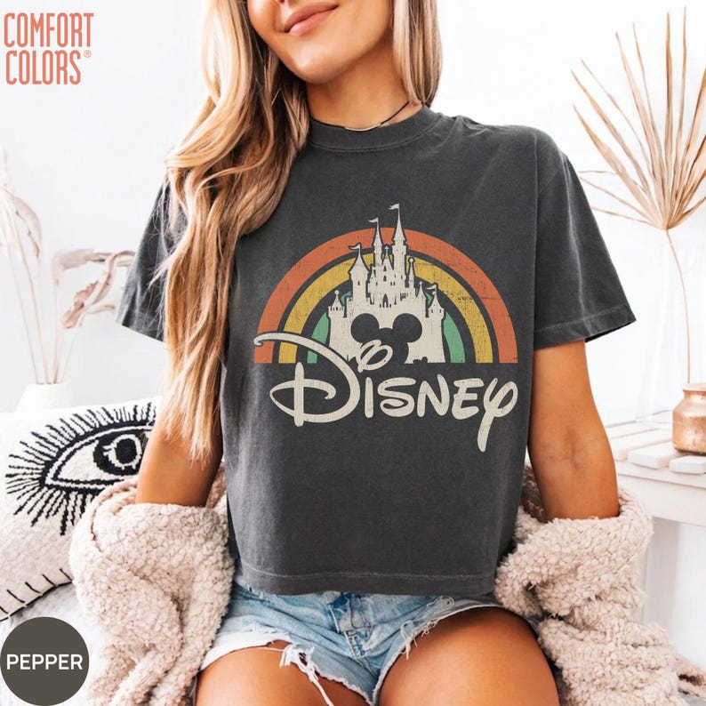 May include: A charcoal gray short-sleeve t-shirt featuring a graphic print of a white castle silhouette centered under a multi-colored rainbow arch. The word Disney is printed in a large white script font below the castle and rainbow graphic.