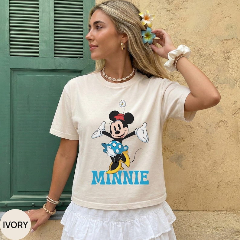 May include: An ivory short-sleeved t-shirt featuring a graphic of the cartoon character Minnie Mouse wearing a red hat and a blue polka-dot skirt. The name MINNIE is printed in large, bold, light blue capital letters below the character graphic.