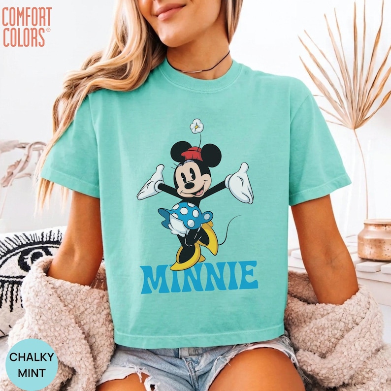 May include: A mint green short-sleeve t-shirt featuring a graphic of Minnie Mouse wearing a red hat and blue polka-dot skirt. The character is depicted with white gloves and yellow shoes. The text MINNIE is printed in blue below the character.