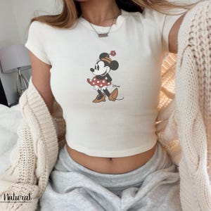 K&ouml;nnte beinhalten: Cremefarbenes Crop-Top mit Minnie Mouse-Grafik. Die Zeichentrickfigur ist mit einem rot-wei&szlig; gepunkteten Rock, braunen Schuhen und einem passenden Hut dargestellt. Das Wort "Disney" ist darunter aufgedruckt.