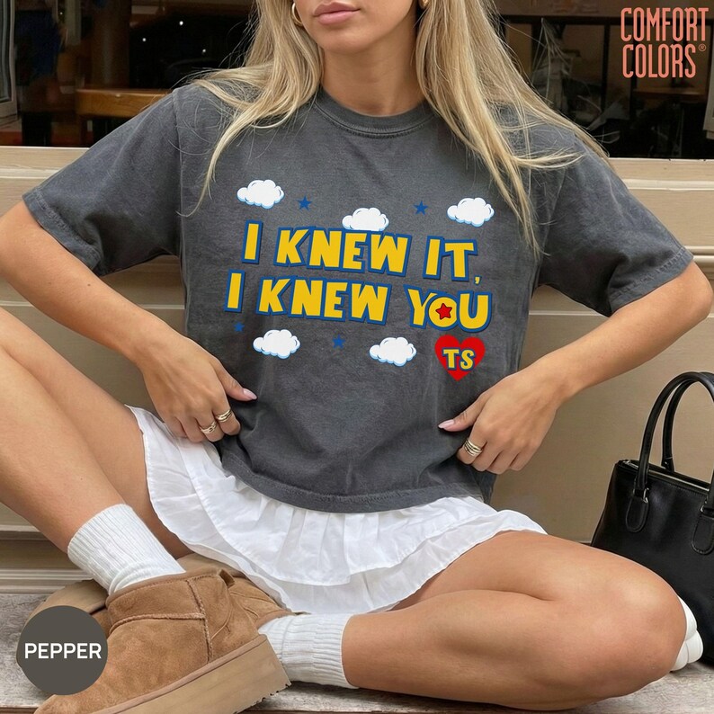 May include: A charcoal gray short-sleeve t-shirt featuring the yellow text I KNEW IT, I KNEW YOU, a red heart with the letters TS, and white cloud graphics. The shirt has a crew neckline and a relaxed fit, with the brand name COMFORT COLORS visible.
