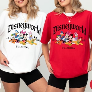 May include: A white t-shirt and a red t-shirt, each featuring a graphic print with the text Disneyworld, est. 1971, and FLORIDA. The design includes illustrations of Mickey Mouse, Minnie Mouse, Donald Duck, Daisy Duck, Goofy, and Pluto.