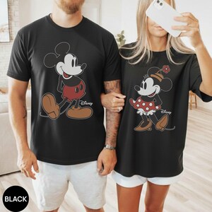 May include: Black t-shirts featuring Mickey and Minnie Mouse. Mickey is in red shorts and brown shoes, and Minnie is in a red polka-dot skirt and brown shoes. Both shirts have the Disney logo.