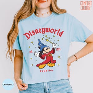 May include: Light blue t-shirt with a graphic of Mickey Mouse as a sorcerer, holding a wand. The design includes the text "Disneyworld" in red, "est. 1971", and "FLORIDA". The shirt is made of a soft material and has short sleeves.