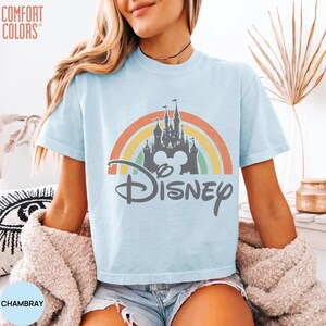 May include: A light blue short-sleeve t-shirt featuring a graphic of a castle silhouette centered over a multi-colored rainbow arch. The word Disney is printed in a dark gray script font below the castle. The text Comfort Colors is visible.