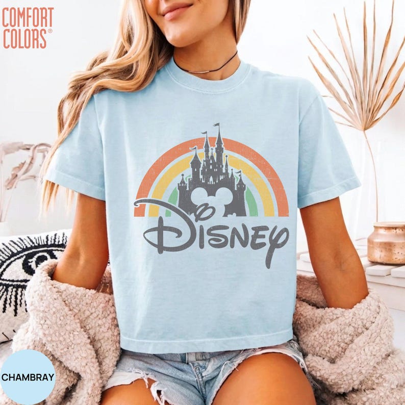 May include: A light blue short-sleeve t-shirt featuring a graphic of a castle silhouette centered over a multi-colored rainbow arch. The word Disney is printed in a dark gray script font below the castle. The text Comfort Colors is visible.