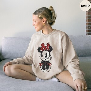 May include: Sand-colored sweatshirt featuring a Minnie Mouse graphic. The graphic depicts Minnie Mouse with a red bow with white polka dots. The sweatshirt is a crew neck style and has long sleeves.