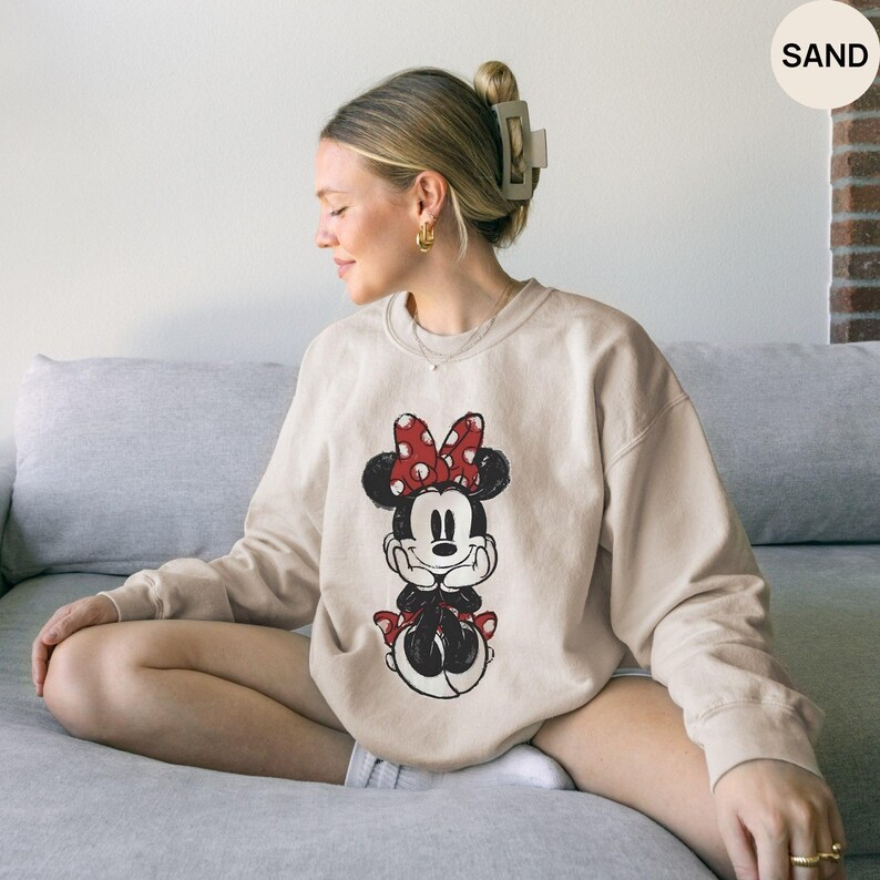 May include: Sand-colored sweatshirt featuring a Minnie Mouse graphic. The graphic depicts Minnie Mouse with a red bow with white polka dots. The sweatshirt is a crew neck style and has long sleeves.