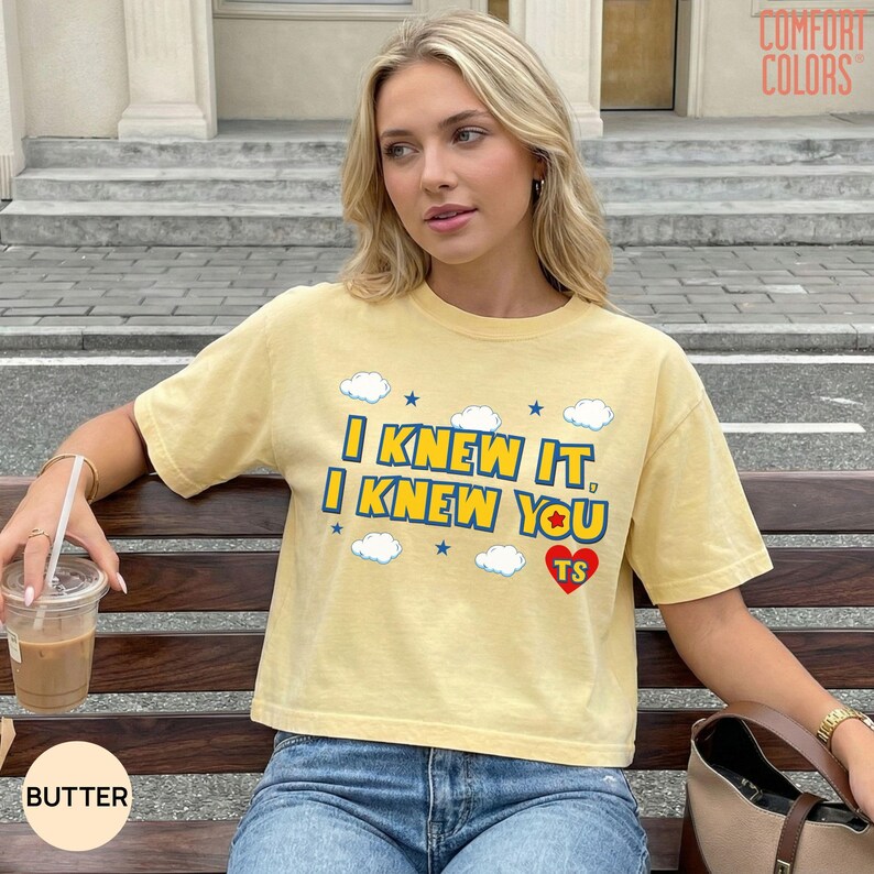 May include: A butter yellow short-sleeve t-shirt featuring the blue text I KNEW IT, I KNEW YOU with small blue stars and white clouds. A red heart with the letters TS is printed below the text. The shirt has a crew neckline and a relaxed fit.