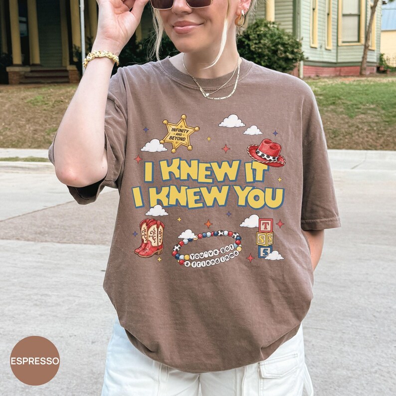 Op de afbeelding: Een espressobruin t-shirt met korte mouwen en een grafische print met een gele sheriffster, een rode cowboyhoed, rode cowboylaarzen, een kralenarmband en alfabetblokken. De tekst luidt I KNEW IT I KNEW YOU, INFINITY AND BEYOND.
