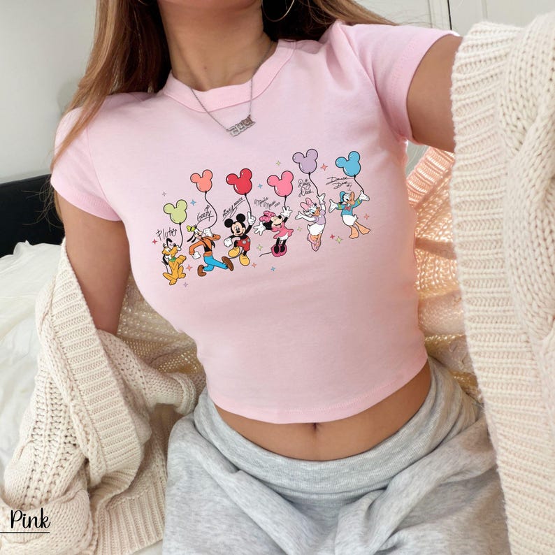 May include: Pink crop top featuring a graphic of Disney characters holding balloons. The characters include Pluto, Goofy, Mickey Mouse, Minnie Mouse, Daisy Duck, and Donald Duck. The word "Pink" is printed at the bottom left.