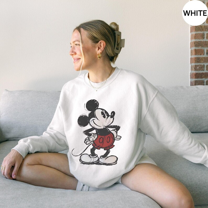 May include: White crewneck sweatshirt featuring a distressed graphic of Mickey Mouse in black, red, and white. The cartoon character is in a standing pose with hands on hips. The sweatshirt is a casual style.