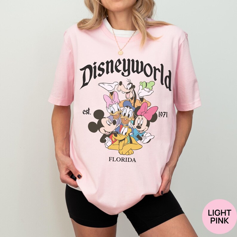 May include: A light pink short-sleeve t-shirt featuring a graphic print of Disney characters including Mickey Mouse, Minnie Mouse, Donald Duck, Daisy Duck, Goofy, and Pluto. The text reads Disneyworld, est. 1971, and FLORIDA in black lettering.