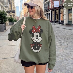 May include: Military green sweatshirt featuring a distressed Minnie Mouse graphic with a red bow. The sweatshirt has a crew neck and long sleeves. The text "MILITARY GREEN" is in the upper right corner.
