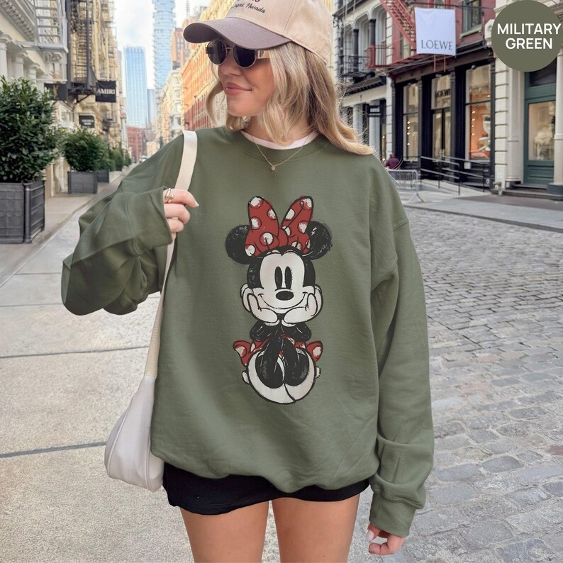 May include: Military green sweatshirt featuring a distressed Minnie Mouse graphic with a red bow. The sweatshirt has a crew neck and long sleeves. The text "MILITARY GREEN" is in the upper right corner.