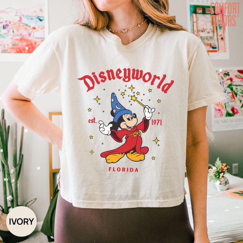 May include: Ivory-colored t-shirt featuring a graphic of Mickey Mouse in a sorcerer's outfit, holding a wand. The text "Disneyworld" is arched above the character, with "est. 1971" and "Florida" below. The shirt is a Comfort Colors brand.